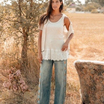 Mulher de blusa branca sem mangas, colete bege e calças de ganga com faixas florais em campo seco próximo a poltrona.