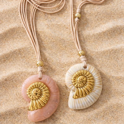 Colares com pendentes em forma de conchas rosa e bege claro com detalhes dourados sobre areia