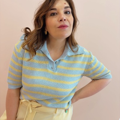 Mulher com camisola de riscas amarelas e azuis e calças amarelas num fundo rosa.