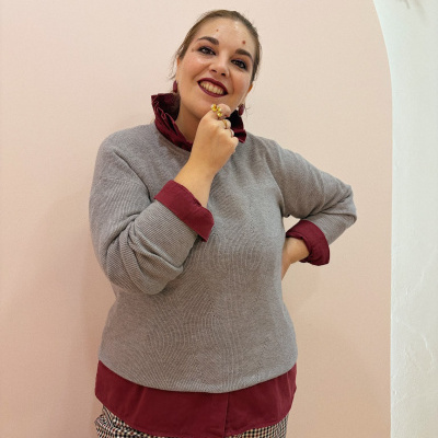 Mulher com blusa cinza, camisa bordô, calças xadrez e acessórios dourados em fundo bege com arco branco