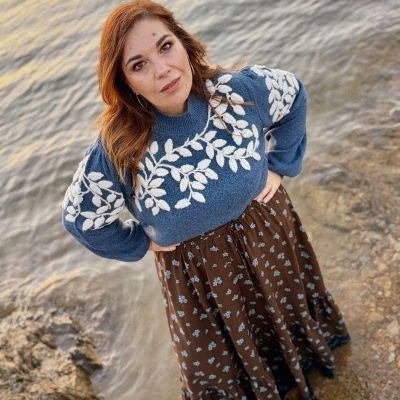 Mulher com suéter azul e saia castanha junto a água e rochas