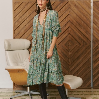 Vestido midi verde com padrão floral e botas pretas em ambiente interior com cadeira e painel de madeira