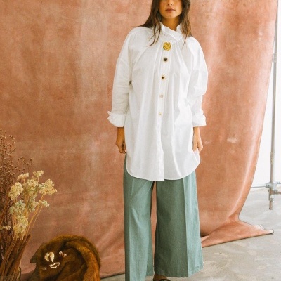 Mulher com camisa branca longa e calças verdes, sapatos bordados, fundo castanho com peles e flores secas