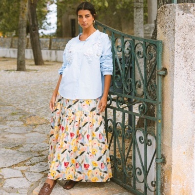 Mulher com camisa azul clara e saia floral comprida encostada num portão de ferro verde