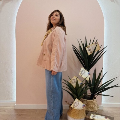 Mulher de perfil com casaco rosa claro e calças de ganga azuis junto a plantas em vasos rústicos num arco branco