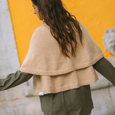 Mulher de costas com poncho bege sobre camisa verde escura, parede amarela e pedra cinzenta ao fundo