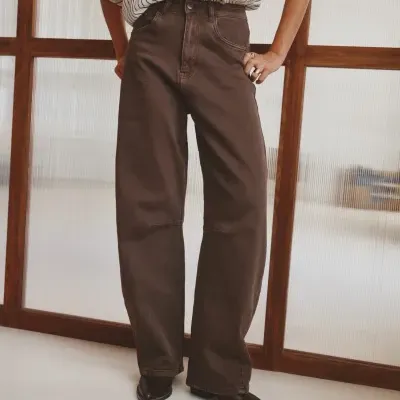 Pessoa com calças de ganga castanhas e camisa riscada branca diante de divisória de madeira.
