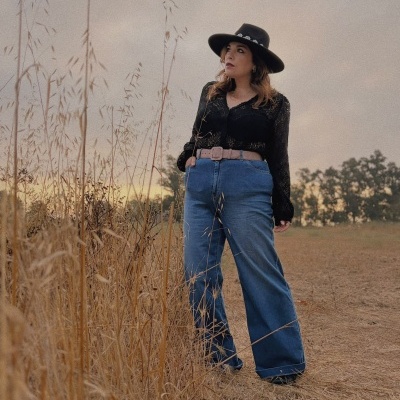 Mulher com blusa preta, calça azul larga, cinto bege e chapéu preto em campo seco.
