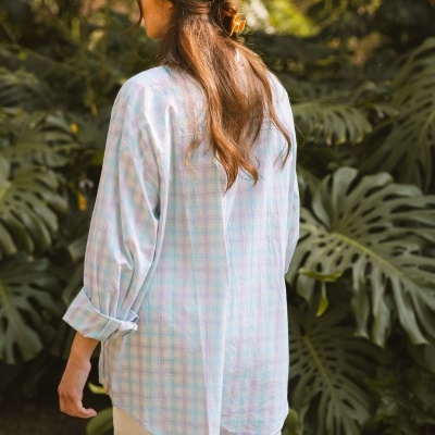 Camisa aos quadrados azul, rosa e branco com calças brancas vista de costas em ambiente exterior com plantas.