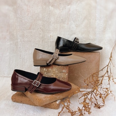 Três pares de sapatos femininos Mary Jane em preto, bege e castanho com fivelas duplas sobre blocos de pedra e fundo de tecido creme.