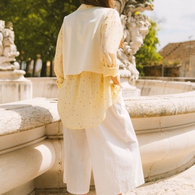 Pessoa de costas com camisa amarela clara, colete branco, calças brancas largas e sapatos marrons junto a fonte de pedra