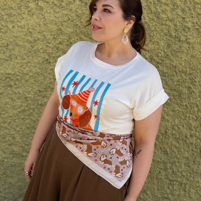 Mulher com t-shirt branca estampada, saia castanha e lenço com padrão de cachorros na cintura diante de parede verde texturizada.