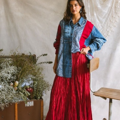 Mulher com camisa que mistura ganga azul, padrão floral e vermelho, e saia longa vermelha enrugada, junto a cadeira de madeira e plantas em baú