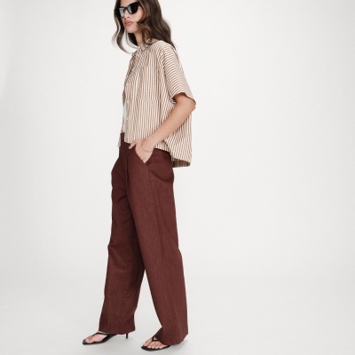 Mulher vestindo calças largas castanhas, camisa curta riscada bege e branca, sandálias pretas e óculos escuros.