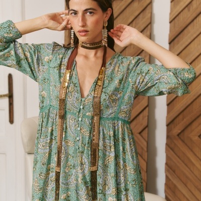Vestido com padrão paisley verde, azul e bege, botões frontais e mangas compridas numa mulher numa sala com parede branca e painel de madeira.