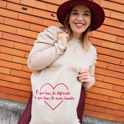 Mala de pano bege com estampa vermelha e texto em português segurada por mulher com chapéu vinho em fundo de parede de tijolos laranja.