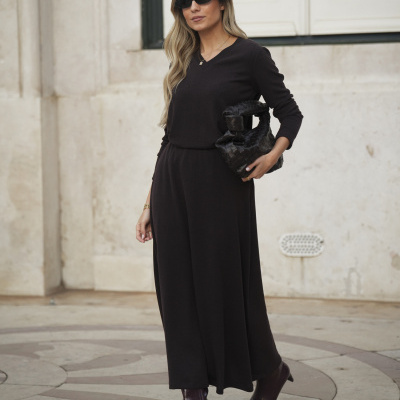 Mulher com vestido preto de manga comprida, botas castanhas, óculos de sol e mala preta