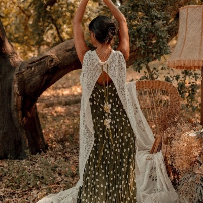 Mulher de costas com vestido verde de bolinhas e capa de malha branca num cenário exterior com cadeira de vime e candeeiro