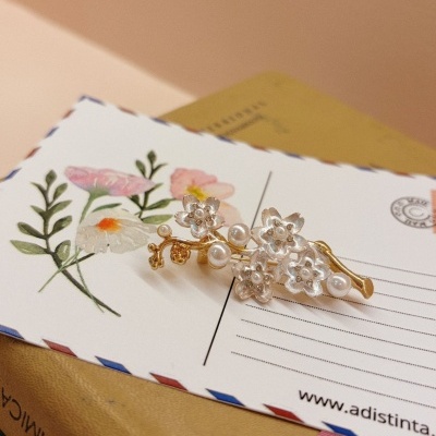 Presilha de cabelo dourada com flores prateadas e pérolas sobre postal floral e livros