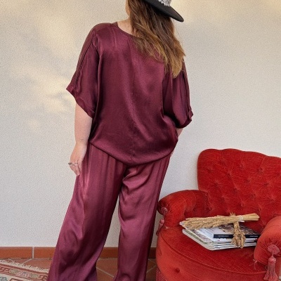 Pessoa de costas com conjunto vinho acetinado e chapéu preto decorado junto a cadeira vermelha acolchoada com revistas.
