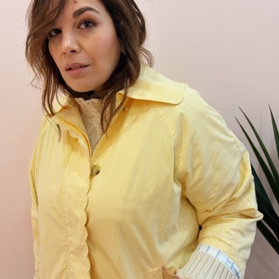 Mulher com casaco amarelo e camisola de malha creme junto a planta verde