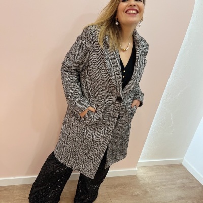 Mulher sorridente com casaco padrão espinha de peixe preto e branco, calças pretas cintilantes e botas pretas em piso de madeira e parede branca e rosa.