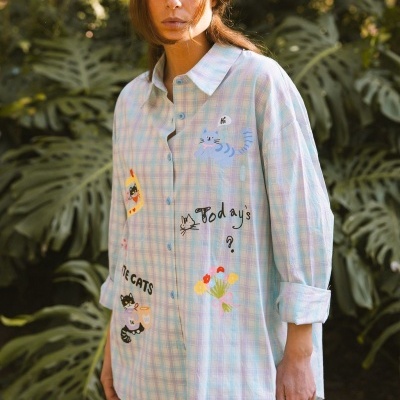 Camisa xadrez azul claro com estampas divertidas de gatos e flores, botões azuis, modelo feminino em ambiente exterior com vegetação
