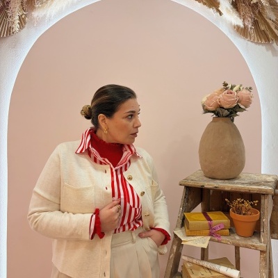 Mulher com camisa vermelha listrada e camisola bege ao lado de escada de madeira com livros, vaso e flores.