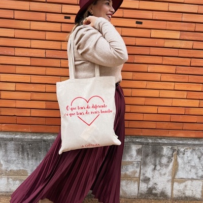 Mulher com saia plissada bordô, botas bege, chapéu bordô e bolsa de pano bege com texto vermelho