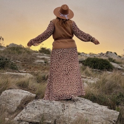 Vista de costas de pessoa com vestido animal print, colete castanho e chapéu castanho em paisagem natural ao pôr do sol