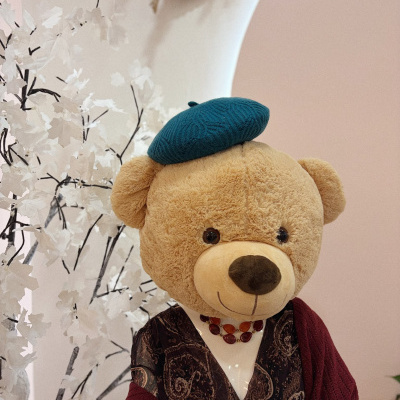 Urso de peluche vestido com boina azul e camisola castanha, em ambiente com decoração branca e parede rosa