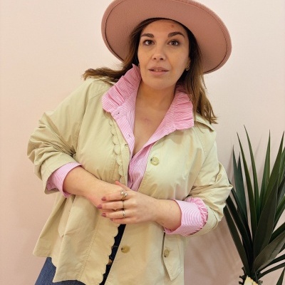 Mulher com chapéu rosa, casaco bege e camisa rosa às riscas, junto a planta com cartão postal.