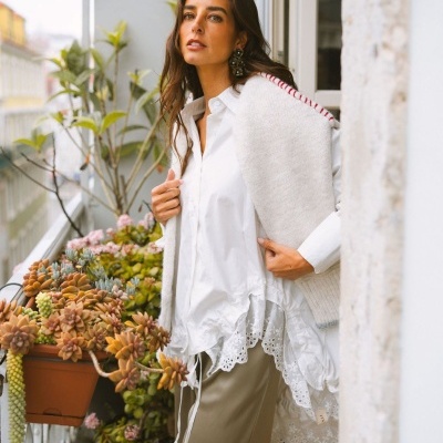 Mulher com camisa branca, colete branco e calças bege numa varanda com plantas