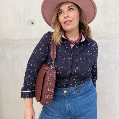 Mulher com chapéu rosa, blusa azul escura com padrões, calças de ganga azuis e mala castanha com detalhes