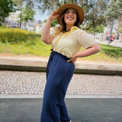 Mulher com camisa amarela e calças azuis largas em ambiente urbano, usando chapéu castanho e sandálias.