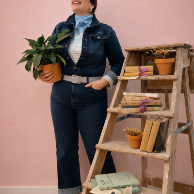 Mulher com roupa de ganga, lenço azul, segura planta verde em vaso castanho, junto a escada de madeira com livros e vasos de plantas, fundo rosa.