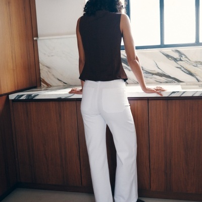 Pessoa de costas com calças brancas e camisola castanha junto a balcão de mármore e móveis em madeira