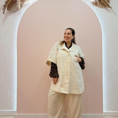 Mulher com casaco bege de manga curta, calças largas bege, camisa preta às bolas brancas e sapatos brancos, em fundo rosa com decoração seca