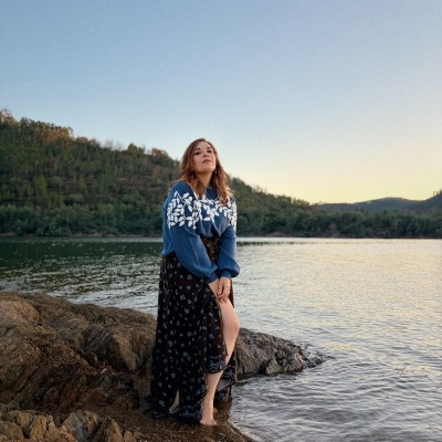 Mulher em rochedos junto à água, veste camisola azul e saia preta com padrão