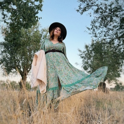 Mulher vestindo vestido verde com padrão cachecol, cinto preto, chapéu preto, casaco bege, em campo seco com árvores e céu azul