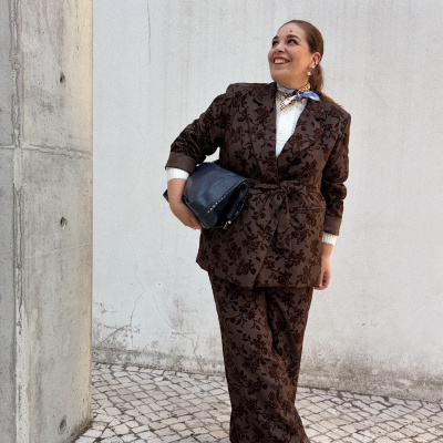 Mulher com fato castanho floral e mala preta em pavimento de pedra e parede branca