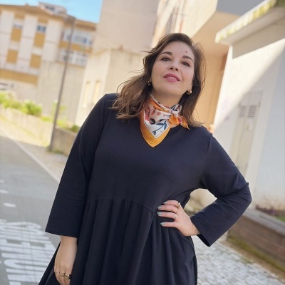 Mulher com vestido preto e lenço colorido ao pescoço em rua urbana.