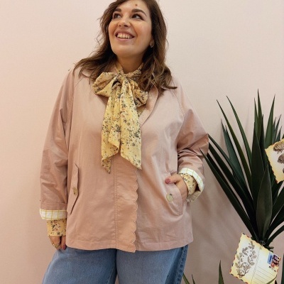 Mulher de casaco rosa, lenço floral amarelo, calças de ganga, ao lado de planta decorada com cartões postais.