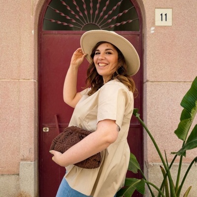 Mulher com chapéu bege e roupa casual com mala castanha perto de porta vermelha