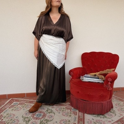 Mulher com vestido castanho e lenço branco na cintura perto de poltrona vermelha