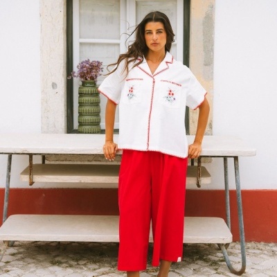 Mulher com camisa branca bordada e calças vermelhas junto a mesa em pedra