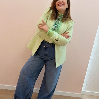 Mulher com blazer verde claro, blusa padrão verde, jeans largos e sapatos verdes em ambiente interior.