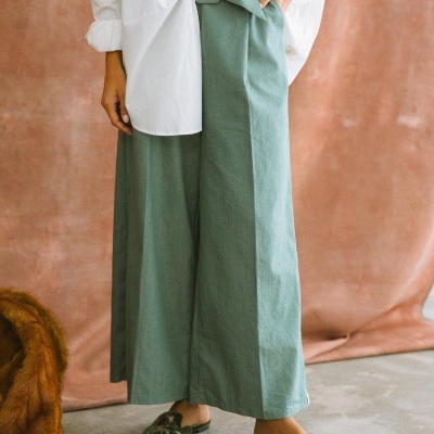 Calças largas verde-água com cinto, camisa branca e mule bordado em fundo castanho.