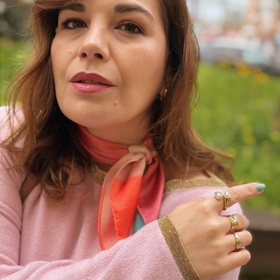 Mulher com blusa de malha rosa, lenço vermelho e anéis dourados.