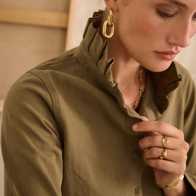 Mulher com brincos, anéis e pulseira dourados e camisa verde-oliva com gola franzida